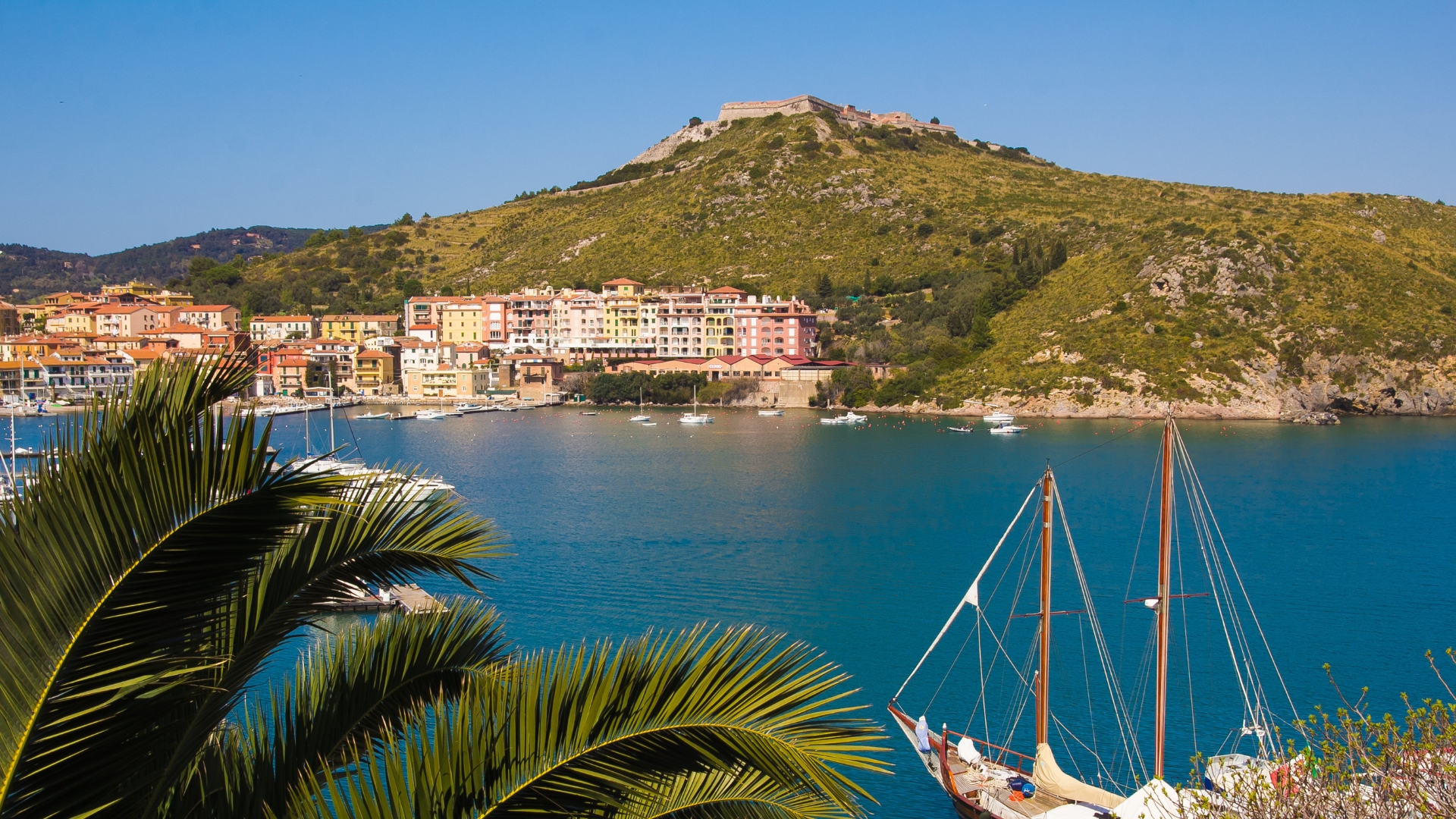 A coastal town with pastel-colored buildings along the waterfront, a green hill rising in the background, calm blue water, a sailing boat in the harbor, and palm fronds in the foreground.