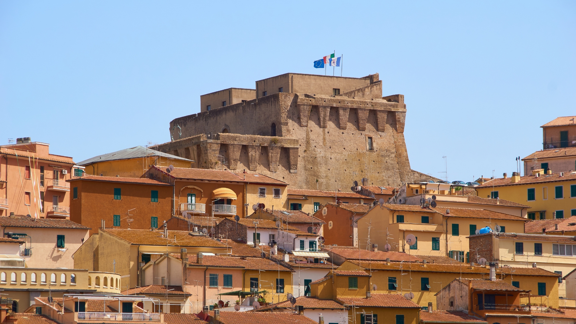 A hillside town dominated by a large stone fortress at the summit, surrounded by terracotta-roofed houses in warm ochre and beige tones, under a bright blue sky.
