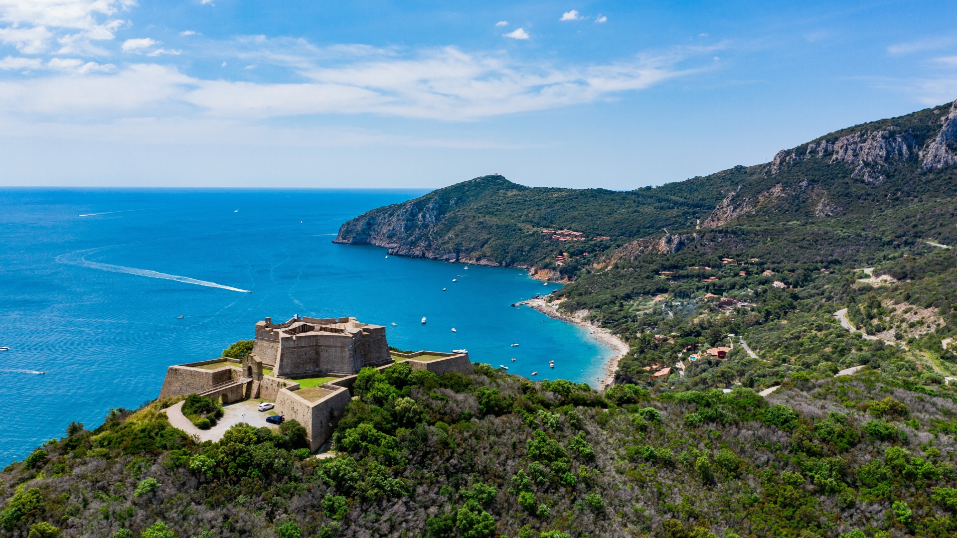 A stone fortress perched on a hill overlooking a deep blue sea, with a curved sandy beach, lush green hills, and a small coastal village along the shore under a bright sky.