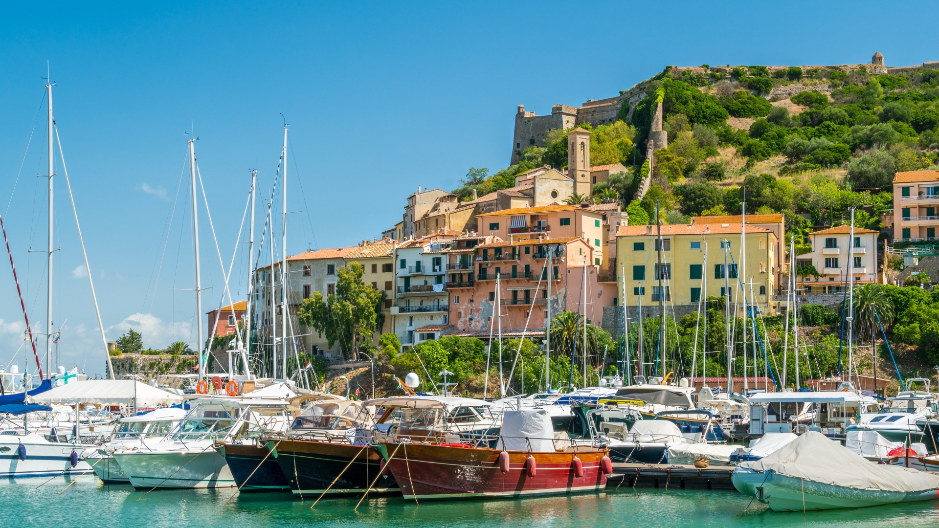 A harbor filled with moored boats and yachts, with a hillside town of pastel-colored houses and a stone fortress perched at the top.