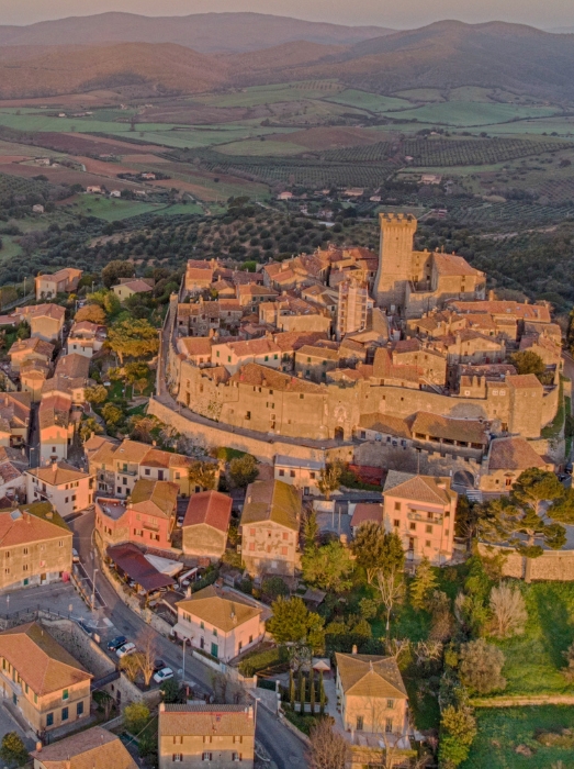Aerial view of a sunlit medieval town perched on a hill, with orange-brown stone buildings, a central castle and surrounding walls, and green countryside beyond.