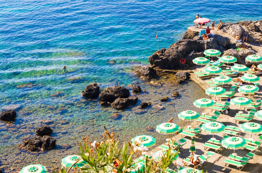 Clear blue sea with a rocky coastline and a stone terrace lined with rows of green and white umbrellas and sun loungers along the water.