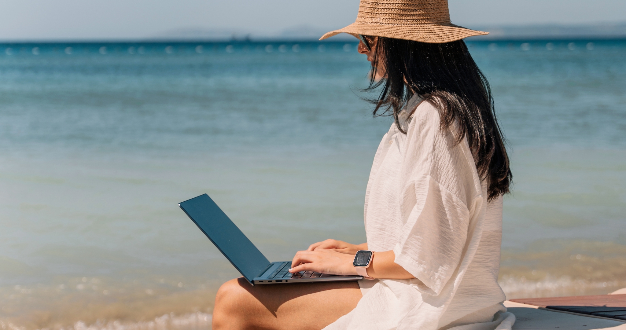 A woman wearing a straw hat and a light dress sits on a sandy beach, using a laptop.