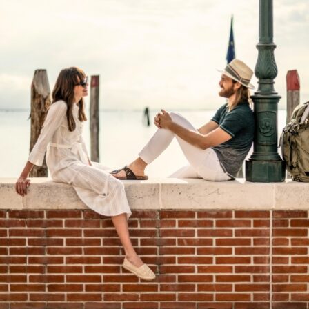 Two people sit on a brick ledge by the water, facing each other and talking. The man wears a hat, a dark t‑shirt, and white pants, while the woman wears a light white dress and sandals. A lamppost and a backpack are nearby, with calm water and posts in the background.