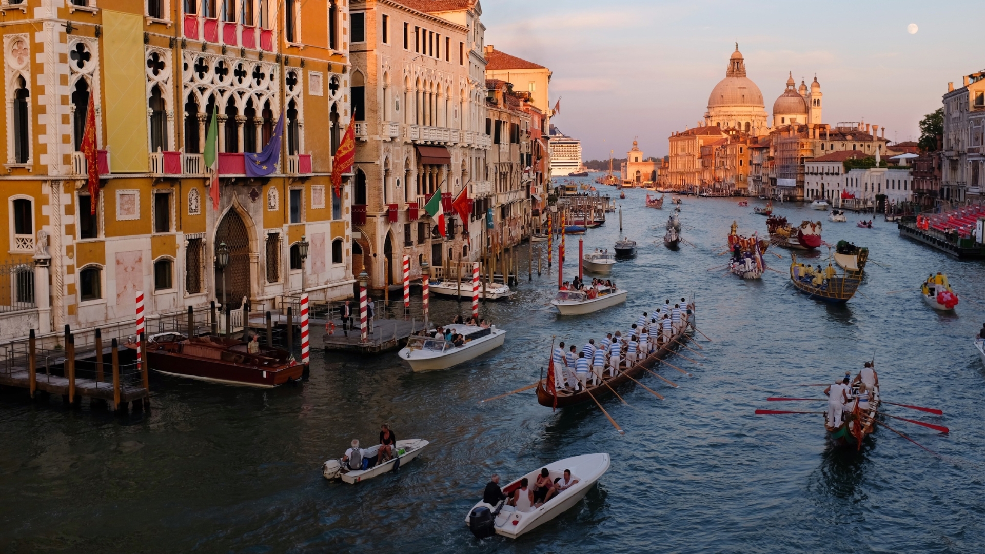 Canale di Venezia con edifici colorati lungo le rive, molte gondole e altre barche sull'acqua, e la Basilica di Santa Maria della Salute sullo sfondo al tramonto.