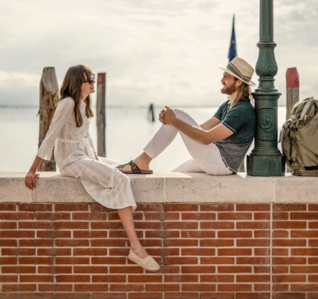 Due persone sedute sul bordo di un muretto di mattoni lungo la riva, con il mare sullo sfondo: una donna in abito bianco e un uomo con cappello e maglietta scura chiacchierano, accanto a una borsa e a uno zaino.