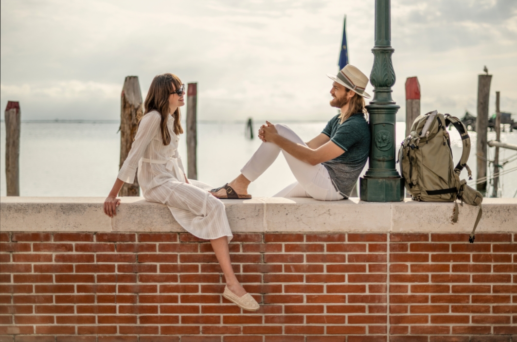 Two people sit on a low brick wall by the water, facing each other and talking. The woman wears a white dress and sunglasses, while the man wears a hat and light clothing. A backpack and a green lamppost sit nearby, with a pier and calm sea in the background.
