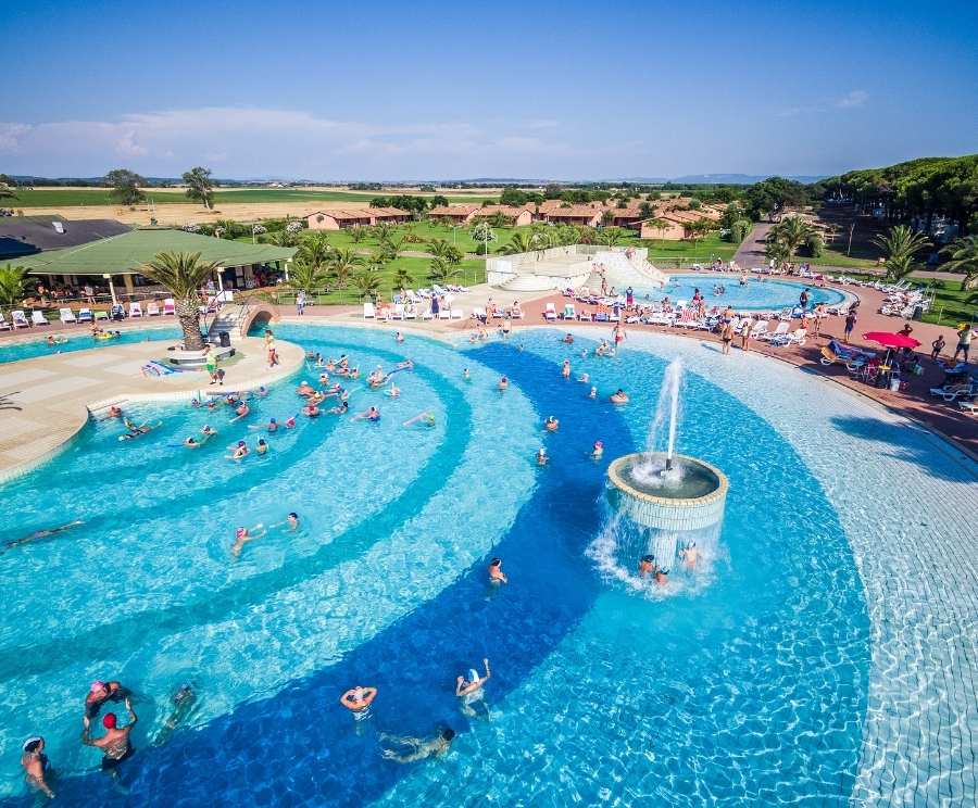 Piscina a forma di anello con fontana centrale, bagnanti che nuotano e si divertono; bordo con lettini e ombrelloni, palme e prato verde sullo sfondo.