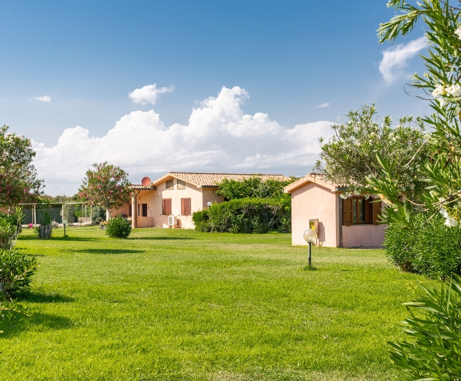 Ampio prato verde con casette basse color rosa pallido, tetti in tegole e finestre con persiane marroni, circondate da alberi e cespugli sotto un cielo azzurro con nuvole bianche.