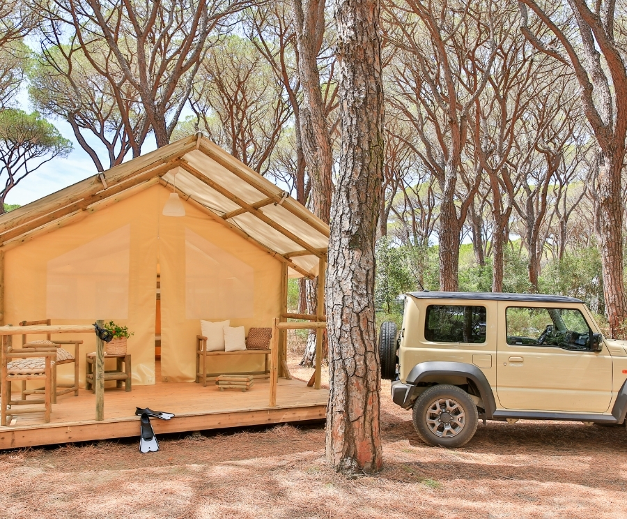 Una tenda in tela beige con struttura in legno è allestita su una piattaforma di legno, con divani e tavolo, circondata da pini; accanto è parcheggiato un fuoristrada beige.