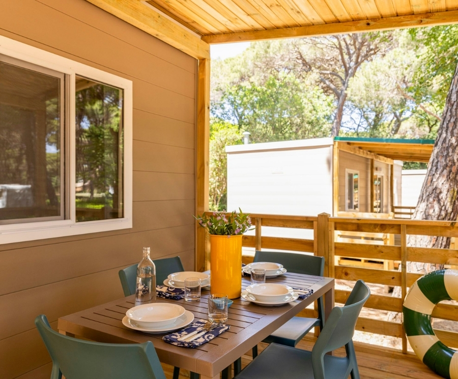 Terrazza esterna con tavolo in legno per quattro, apparecchiato con piatti bianchi, bicchieri e tovagliette; al centro un vaso giallo con fiori, una bottiglia d'acqua e sedie blu. Sullo sfondo una parete di legno, un piccolo edificio bianco e alberi, con un salvagente appeso alla ringhiera.