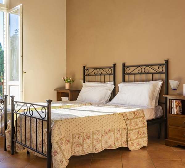 Camera da letto con due letti in ferro battuto, coperte beige a motivo floreale e cuscini bianchi. Due comodini ai lati con lampade: sul comodino sinistro una pianta, sul destro una piccola libreria. Pavimento in piastrelle di terracotta e pareti color sabbia; una porta-finestra aperta lascia entrare la luce dall'esterno verde.