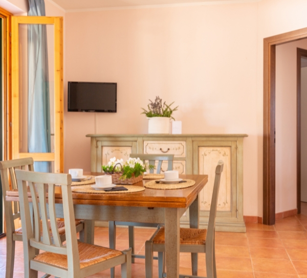 Sala da pranzo accogliente con tavolo in legno e quattro sedie, apparecchiatura tavola con piatti e tazze, una credenza verde pallido sullo sfondo con una pianta, e un piccolo TV a parete; pavimento in piastrelle color terracotta e una porta a sinistra.