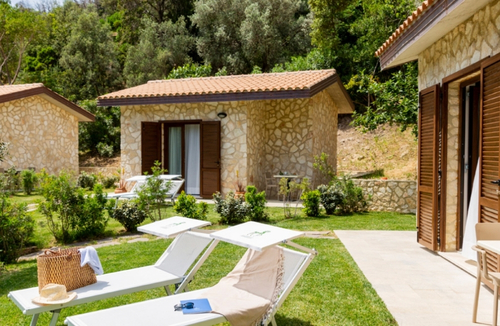 Due casette in pietra con tetto in tegole, giardino soleggiato; sedie a sdraio bianche sul prato, una borsa di paglia e un cappello poggiati su una sedia, e porte in legno delle abitazioni.
