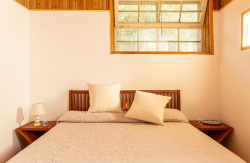 Camera da letto luminosa con letto matrimoniale coperto da coperta beige, due cuscini chiari sulla testiera in legno, comodini ai lati con lampade, finestra in legno sopra il letto e pareti bianche con soffitto in legno.
