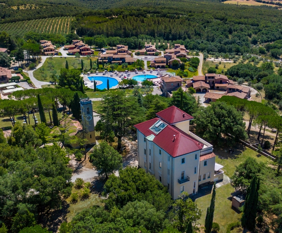 Vista aerea di un resort circondato da alberi e colline: edifici con tetti rossi, una torre, una grande piscina azzurra circondata da lettini e villette disposte lungo strade serpeggianti.