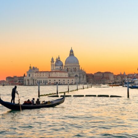 Una gondola con passeggeri e il gondoliere al timone naviga sul canale di Venezia al tramonto, con la Basilica di Santa Maria della Salute sullo sfondo e un cielo arancione.