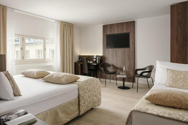 Camera d'albergo luminosa con due letti separati, coperte beige e cuscini, zona studio con scrivania e sedia, TV montata su pannello di legno, due poltrone nere e un tavolino rotondo in vetro, grande finestra con tende chiare.
