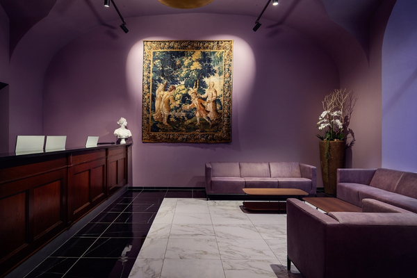 Interno di una hall elegante con pareti viola, un dipinto incorniciato al centro della parete, un bancone in legno a sinistra, divani e poltrone viola, pavimento in marmo bianco e nero, e un grande vaso di piante sulla destra.