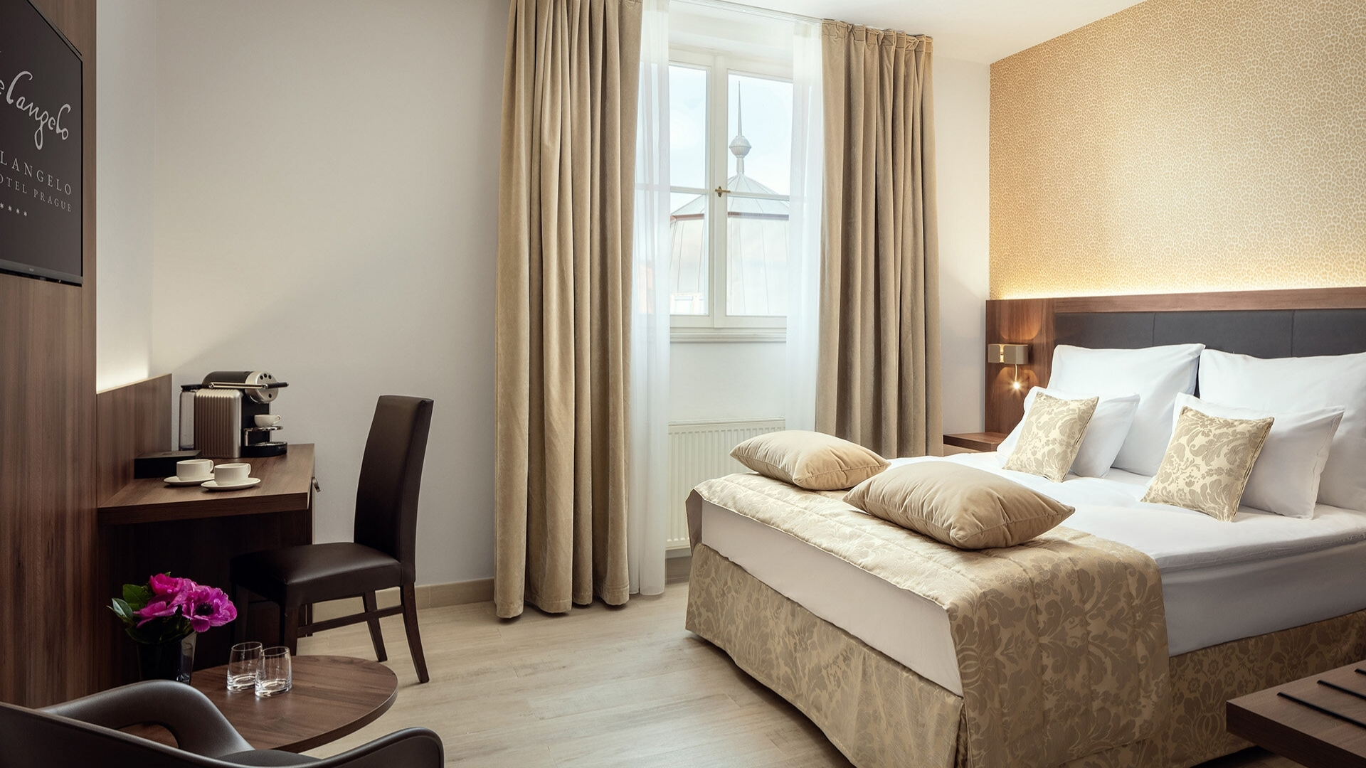 Camera d'albergo luminosa e accogliente: letto matrimoniale bianco e crema con molti cuscini beige e testiera imbottita, comodini con lampade, tende beige e finestra. In primo piano tavolino rotondo con bicchieri e un vaso di fiori rosa; accanto una sedia. Sullo sfondo scrivania con macchina da caffè e altra sedia.