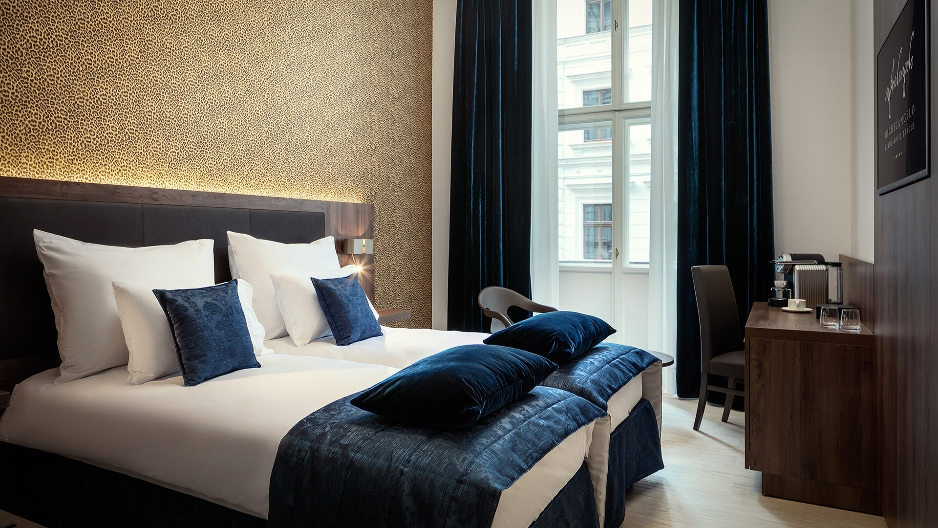 Camera d'albergo moderna con letto matrimoniale bianco, cuscini e coperta blu scuro, parete dorata alle spalle, tende blu scuro e grande finestra; scrivania con set da tè e tazze sulla destra.