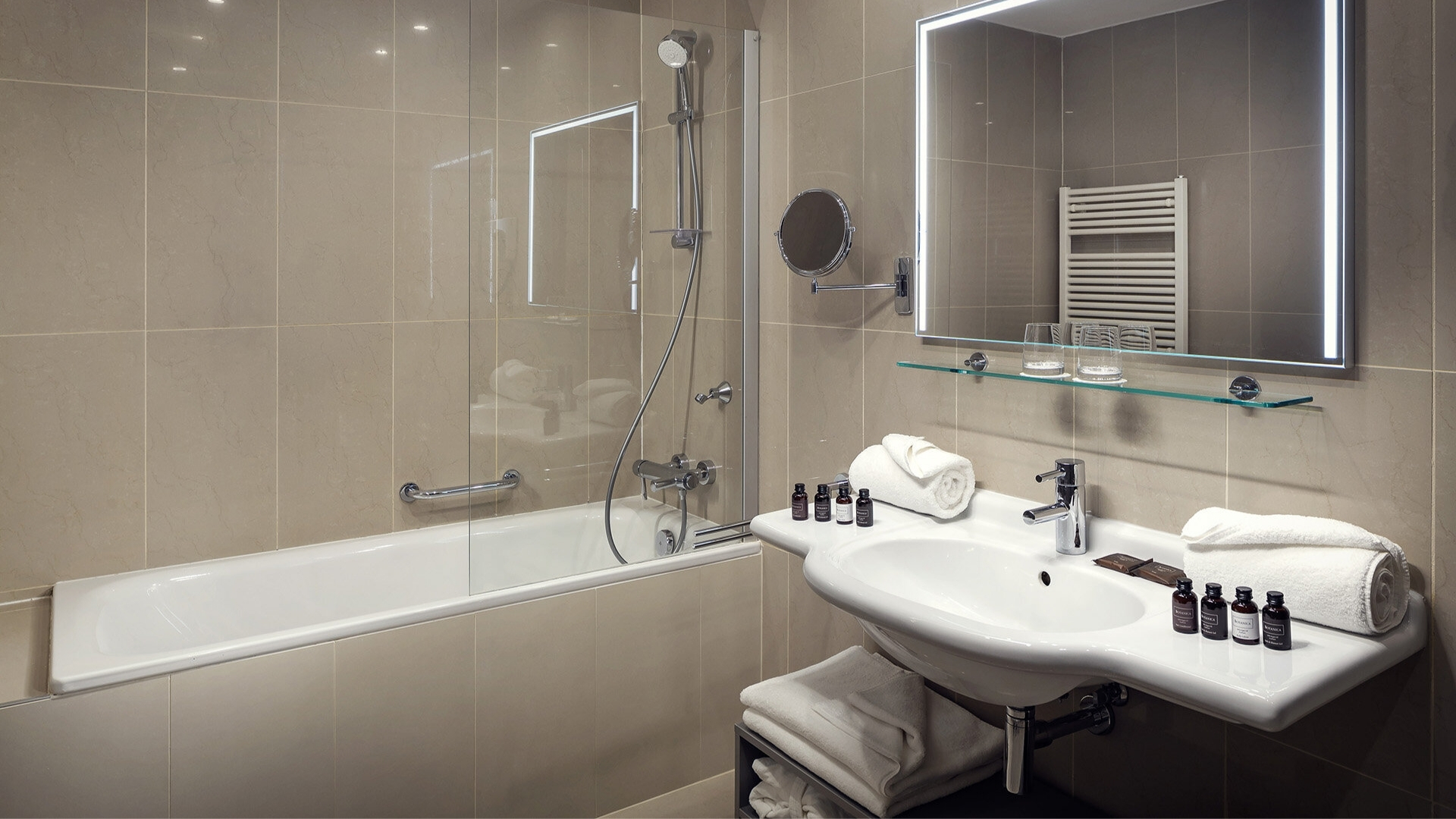 Bagno moderno con vasca da bagno e cabina doccia in vetro, grande lavabo bianco con rubinetto cromato, asciugamani piegati su una mensola e sotto il lavabo, una serie di piccoli flaconi di prodotti da bagno sul piano. Uno specchio largo con cornice luminosa è posto sopra il lavandino.