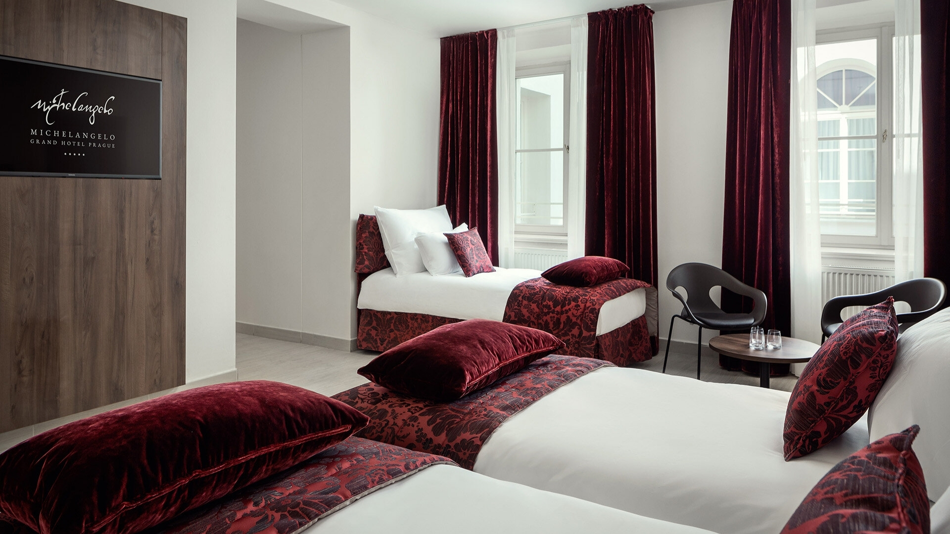 Camera d'albergo luminosa con due letti singoli coperti da biancheria bianca e cuscini rossi, tende bordeaux, zona relax con sedie nere e un tavolino, ampia finestra che lascia entrare la luce.