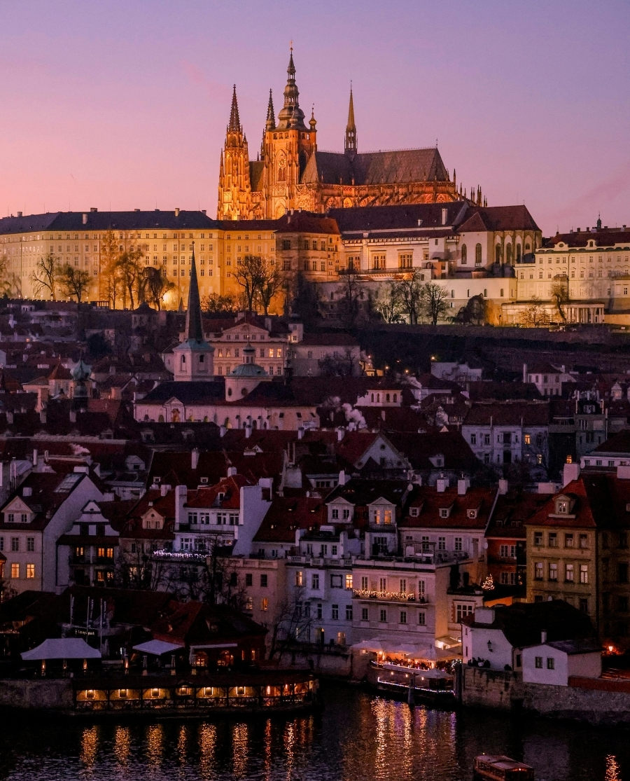 Vista serale di una città europea con tetti rossi, dominata da un grande castello e una cattedrale dalle guglie situati sulla collina; sullo sfondo si intravede un fiume e le luci delle abitazioni.