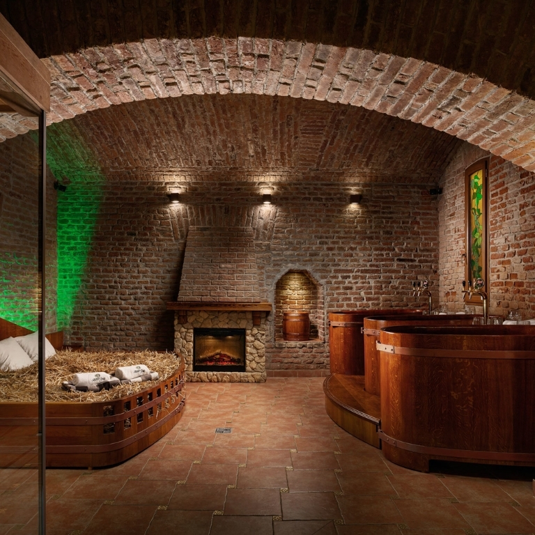 Interno di una cantina/spa in mattoni a volta: camino di pietra acceso al centro, nicchia ad arco sullo sfondo, grandi tinozze rotonde in legno sul lato destro, pavimento in piastrelle rosse e illuminazione soffusa.