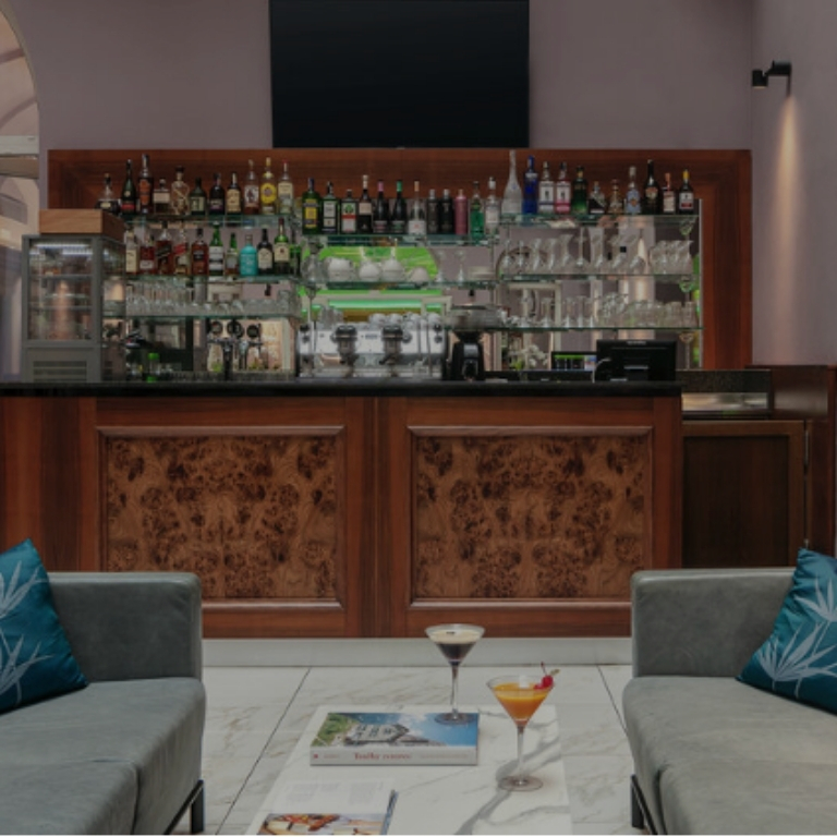 Interno di un bar elegante: bancone in legno con bottiglie e bicchieri sugli scaffali, divani grigi con cuscini blu ai lati e un tavolino in marmo al centro con riviste e due cocktail.