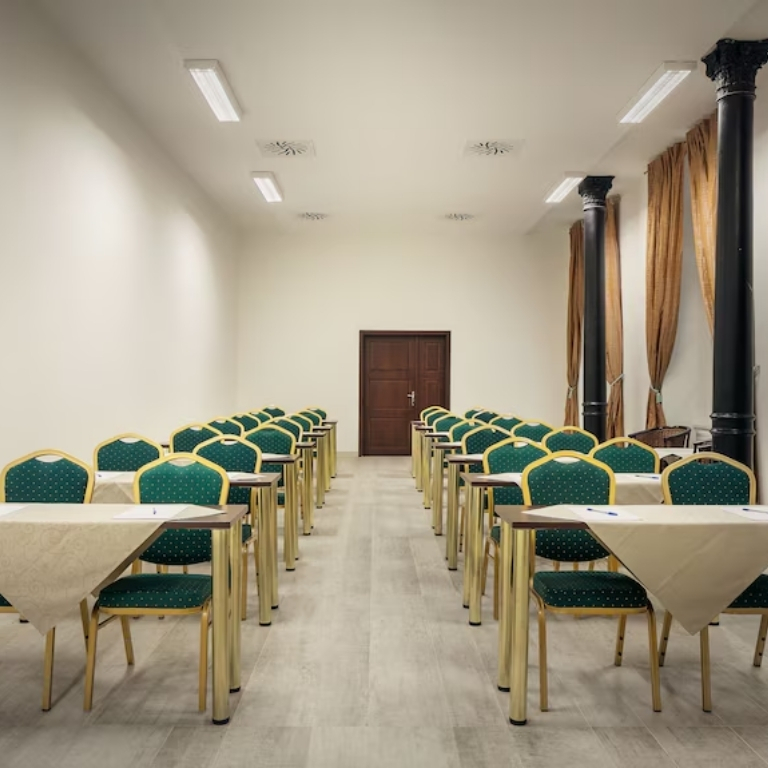 Una sala conferenze luminosa con file di sedie verdi con cornice dorata disposte su entrambi i lati di un corridoio, tavoli coperti da tovaglie bianche, una porta in fondo, colonne nere e tende marroni lungo il lato destro.