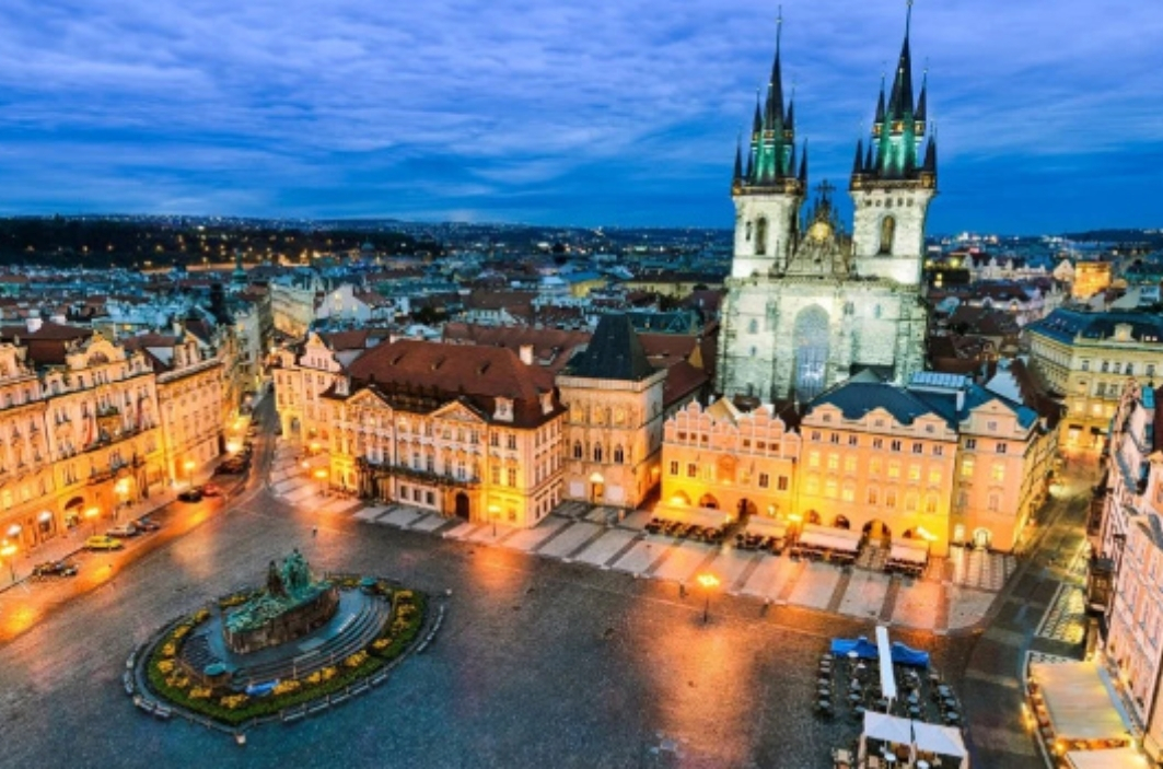 Piazza della Città Vecchia di Praga al crepuscolo, con una fontana circolare al centro e edifici colorati attorno; sullo sfondo si staglia una grande chiesa gotica a due torri.