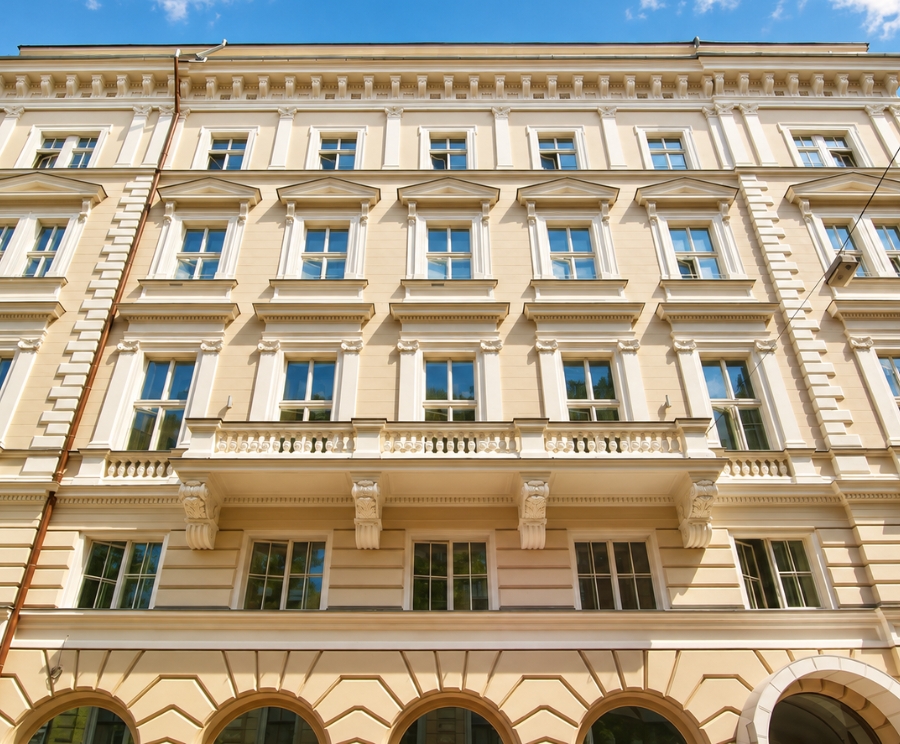 Edificio storico di facciata crema con molte finestre ornate, un balcone centrale e archi al piano terra contro un cielo azzurro.