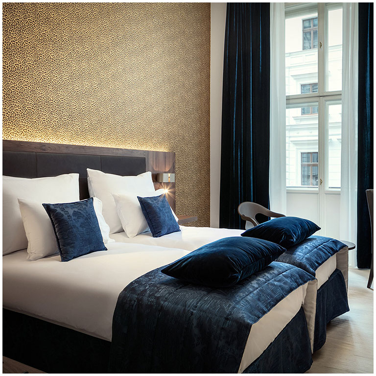 Camera d'albergo con letto matrimoniale coperto da lenzuola bianche, cuscini blu scuro e una coperta blu, testiera imbottita, parete dorata a motivo testurizzato sullo sfondo, grande finestra con tende scure che lasciano entrare la luce.