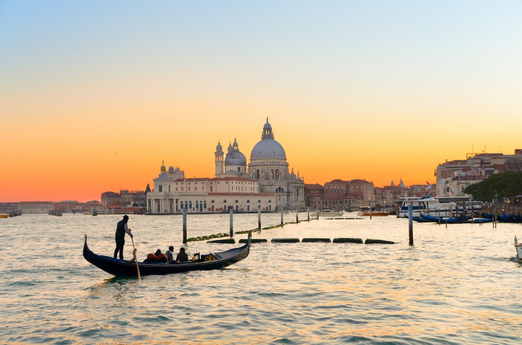 A gondola carrying passengers on a Venetian canal at sunset, with the Santa Maria della Salute basilica and other historic buildings in the background.