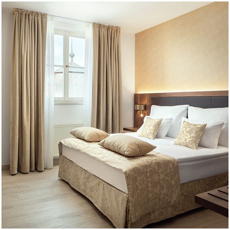 Camera da letto luminosa ed elegante con letto matrimoniale, cuscini beige e copriletto jacquard, lenzuola bianche; tende beige, parete rivestita e comodini con lampade laterali.