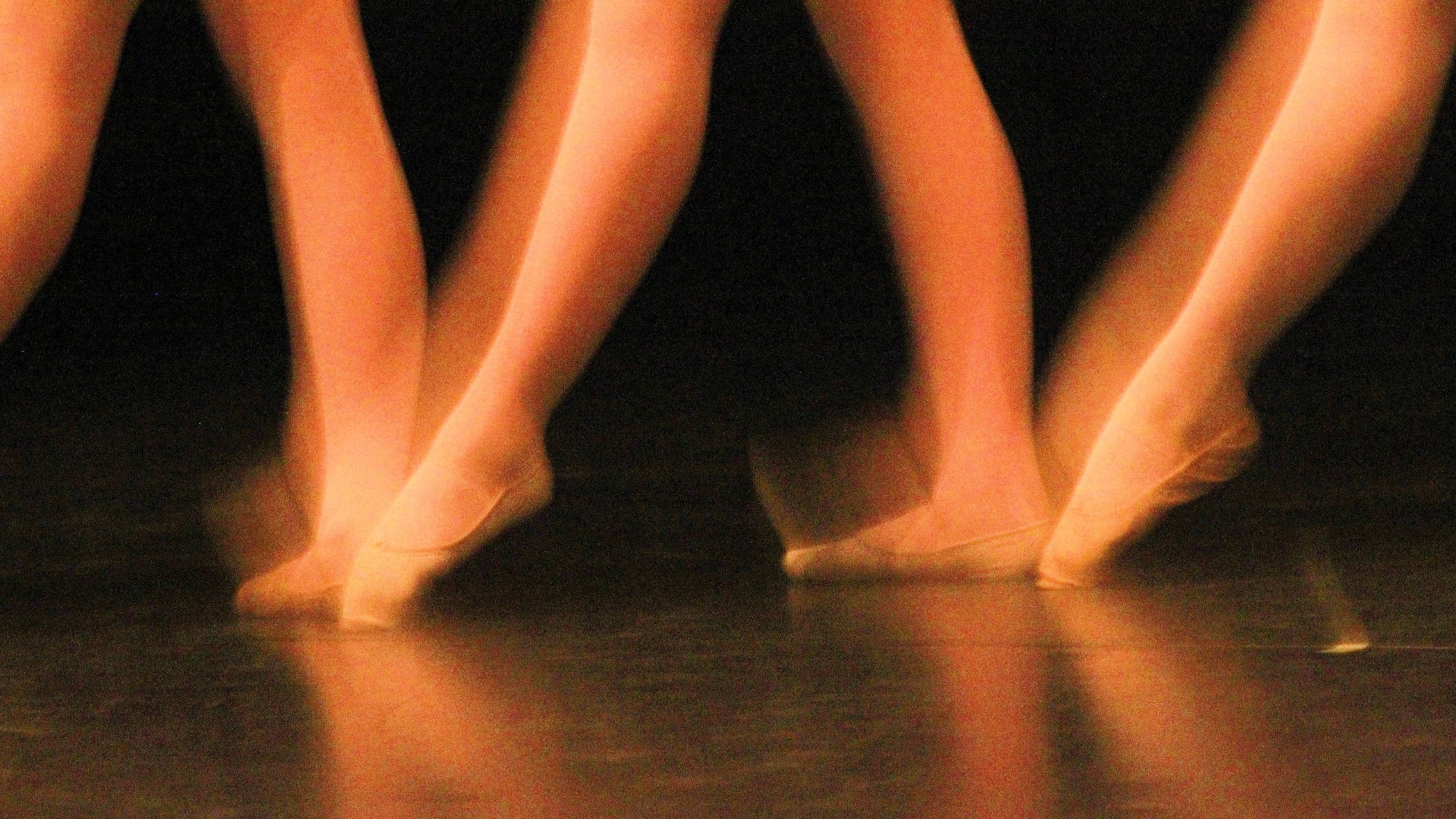 Several ballet dancers on pointe, wearing pointe shoes, standing on a reflective stage illuminated by warm orange light.