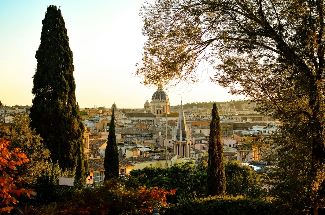 A cityscape at sunset viewed from a hillside, with a domed church in the distance framed by tall cypress trees and autumn-colored foliage.