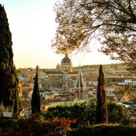 A panoramic view of an Italian city at sunset, featuring a large domed church at the center, tall bell towers, and cypress trees in the foreground.