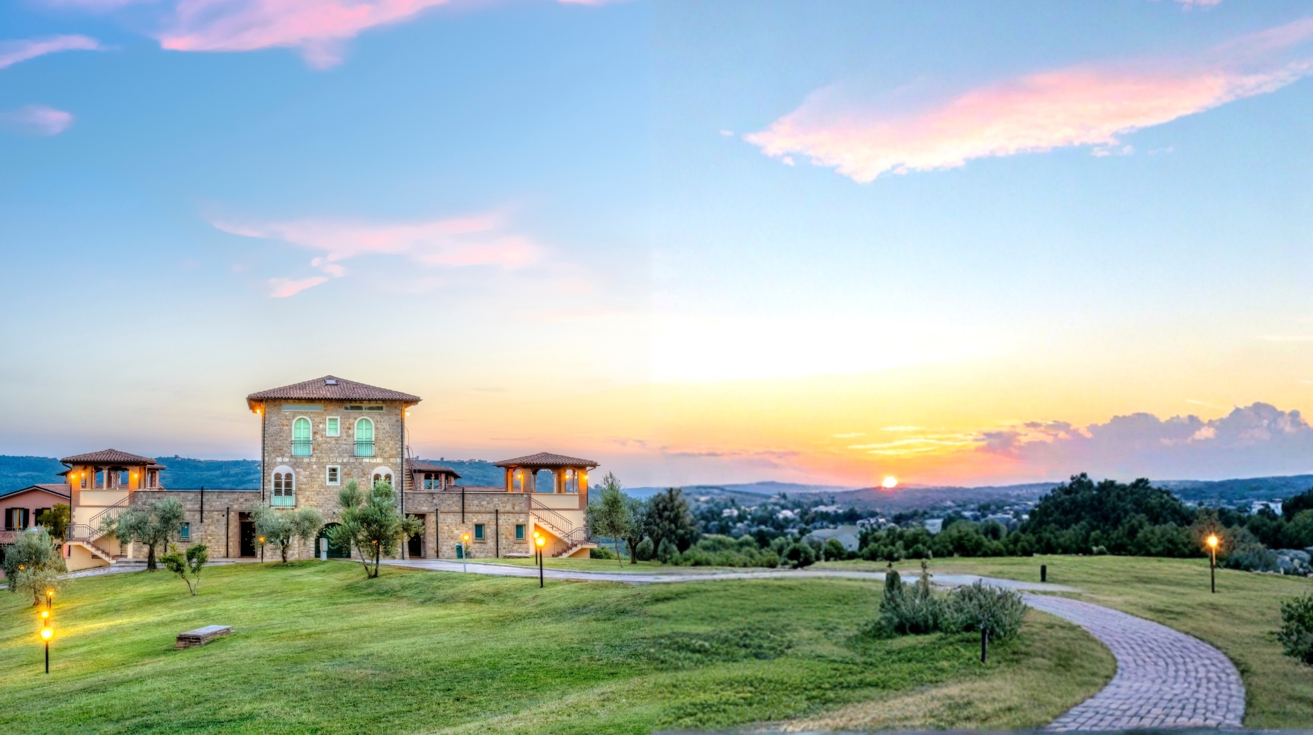 Vista panoramica di un prato verde con una villa in pietra a piú ali, una strada lastricata che curva e lampioni accesi, al tramonto con cielo arancione e nuvole.