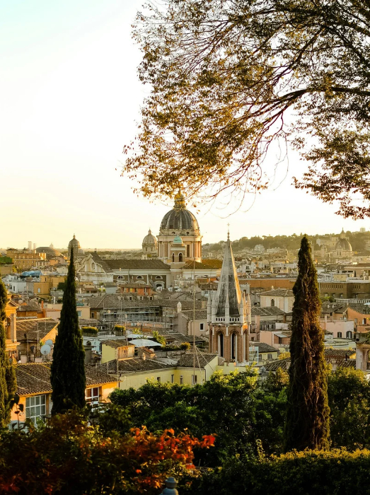 Panoramablick auf eine mediterrane Stadt bei Sonnenuntergang, mit einer markanten Kuppel und Kirchtürmen; im Vordergrund stehen Bäume und Zypressen.