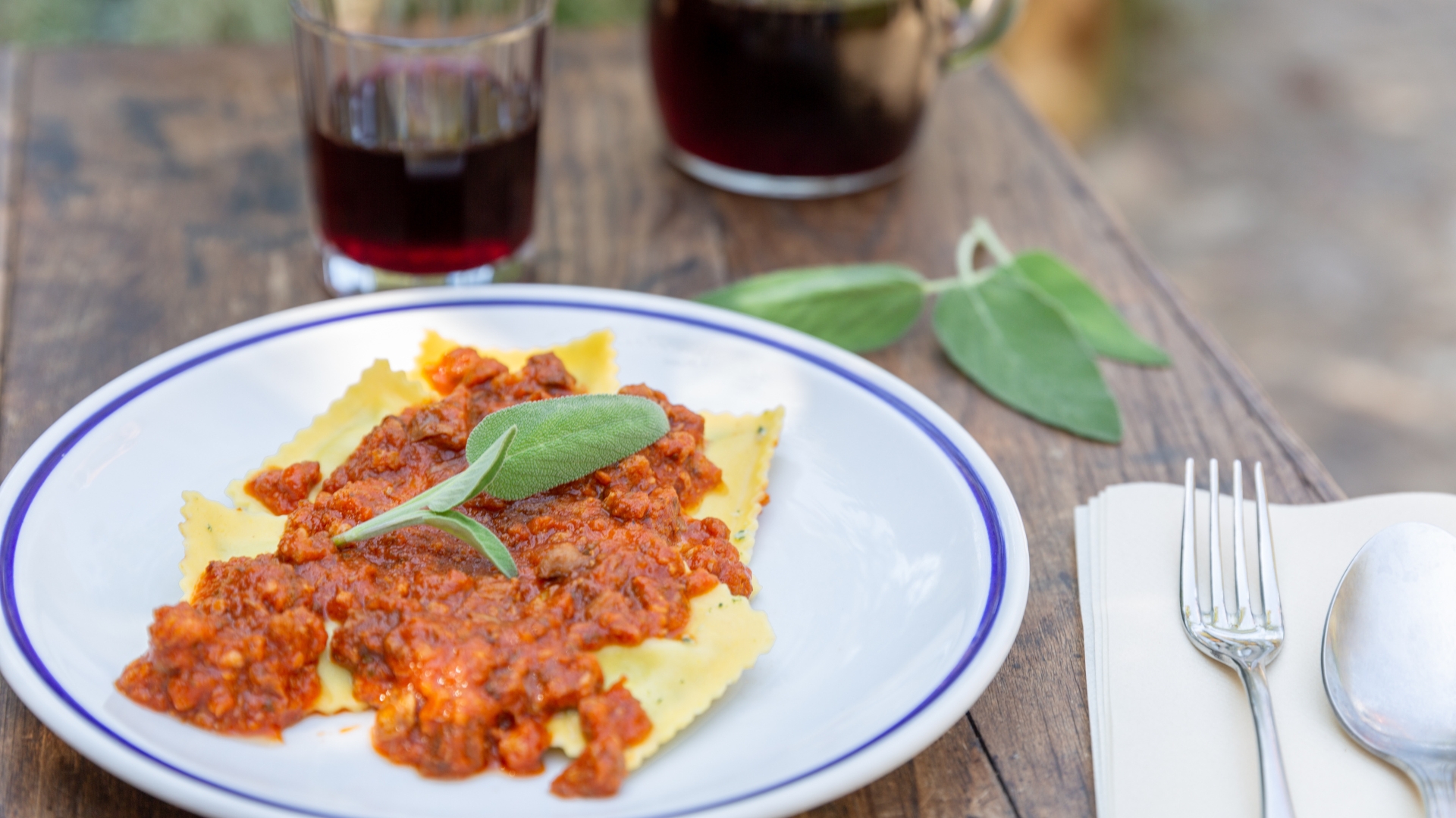 Piatto di pasta fresca condità con ragù di pomodoro, guarnita con foglie di salvia, posto su tavolo di legno; sullo sfondo ci sono due bicchieri di vino rosso e posate accanto a una tovaglietta.