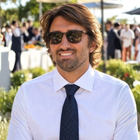 Benjamin Francorsi with brown hair and a light beard wearing sunglasses, a white dress shirt, and a dark tie, smiling at an outdoor event with blurred guests and greenery in the background.