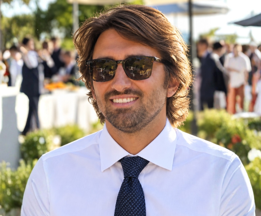 A smiling Benjamin Francorsi with brown hair and a beard wearing sunglasses, a white dress shirt and a dark tie, at an outdoor event with people and greenery in the blurred background.