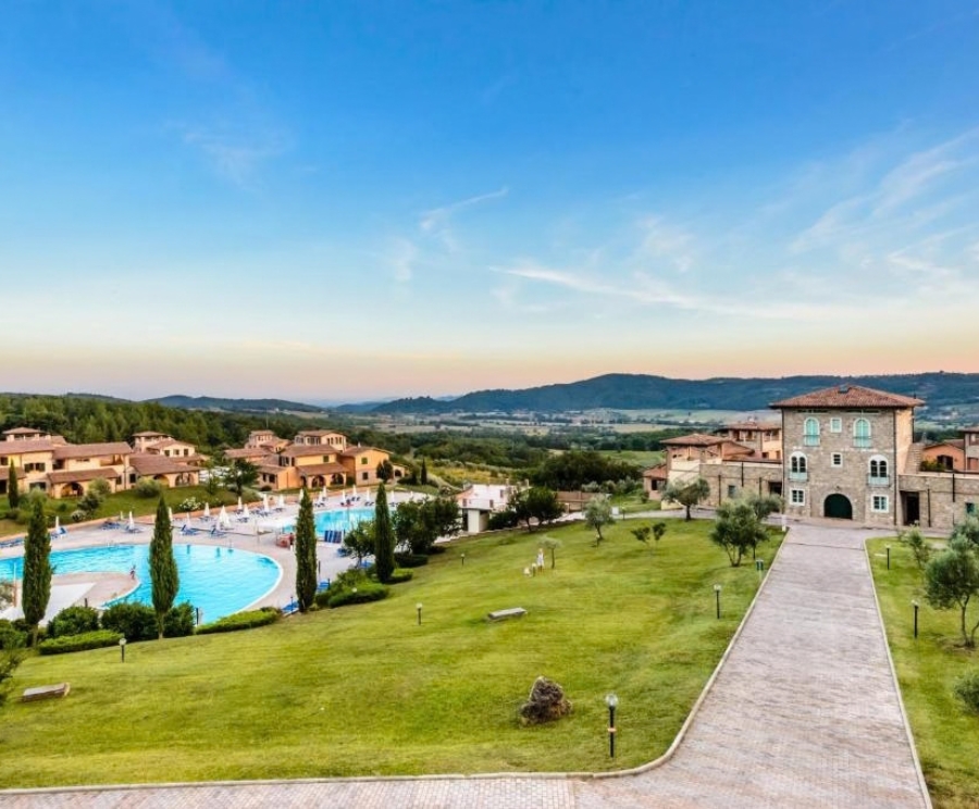 A scenic hillside resort with a curved blue swimming pool on the left, rows of sun loungers and umbrellas, and small beige buildings with orange-tiled roofs around lush green lawns. A stone walkway leads to a large two-story stone mansion on the right, with distant hills and a clear blue sky in the background.