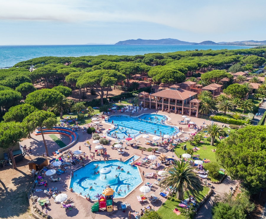 Aerial view of a seaside resort with two swimming pools surrounded by sun loungers and umbrellas, a central building, lush green pine trees, and the sea with distant hills in the background.